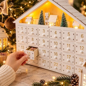 May include: A white wooden house-shaped advent calendar with twenty-four numbered drawers. The top features miniature buildings, green bottle brush trees, and warm interior lighting. Drawer number fourteen is partially open, revealing a small gift.