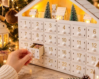 Könnte beinhalten: Ein weißer Adventskalender aus Holz in Hausform mit vierundzwanzig nummerierten Schubladen. Oben befinden sich Miniaturgebäude, grüne Flaschenbürstenbäume und eine warme Beleuchtung. Die Schublade Nummer vierzehn ist leicht geöffnet und zeigt ein kleines Geschenk.