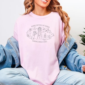 Puede incluir: Sudadera rosa claro con un dibujo lineal negro de una persona rodeada de gatos. El texto en la camiseta dice "BOARD CERTIFIED CAT HERDER" y "EVIDENCE-BASED CHAOS". La persona lleva una chaqueta vaquera.