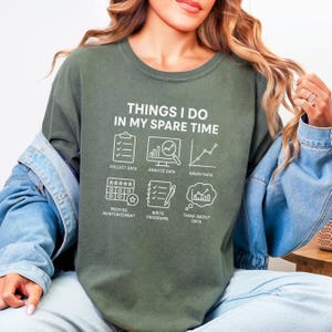 Puede incluir: Sudadera verde oliva con el texto "THINGS I DO IN MY SPARE TIME" e iconos relacionados con datos. Los iconos incluyen una portapapeles, una pantalla de ordenador con una lupa, un gráfico, estrellas, un lápiz y una burbuja de pensamiento.