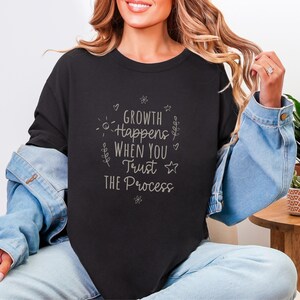 May include: Black crew neck sweatshirt with the text "Growth Happens When You Trust The Process" in a light gray script. The design includes small floral and star accents. The sweatshirt is worn with a denim jacket and blue jeans.