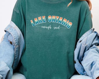 May include: A dark teal crewneck t-shirt featuring the text LAKE ONTARIO in a retro-style arched font with a gradient of orange, yellow, and light blue, followed by the cursive script text enough said, layered under a light blue denim jacket.