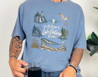 May include: A dusty blue t-shirt featuring a graphic print of Lake Ontario with a lighthouse, pine trees, a flying seagull, and a stack of stones. The text reads, It will always be Lake Ontario to me, centered among the various nature illustrations.