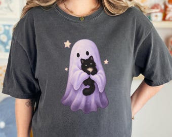 May include: A charcoal gray short-sleeve t-shirt featuring a graphic print of a light purple ghost holding a fluffy black cat. The ghost is surrounded by small, pale yellow stars. The shirt has a crew neckline and a relaxed, boxy fit.