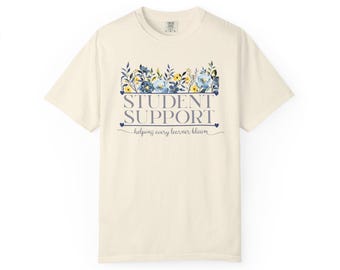 May include: A cream-colored short-sleeve t-shirt featuring a graphic print of blue and yellow flowers above the text "STUDENT SUPPORT" in a dark blue serif font, with the script text "helping every learner bloom" centered below the words.
