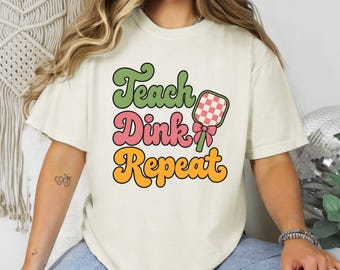 May include: A cream-colored short-sleeve t-shirt featuring the text Teach Dink Repeat in green, pink, and yellow. The design includes a graphic of a checkered pickleball paddle with a pink bow. The shirt is paired with blue denim jeans.