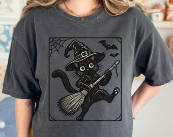 May include: A charcoal gray short-sleeve t-shirt featuring a black graphic print of a black cat wearing a witch hat and riding a broomstick. The illustration includes a spider web in the top left corner and two small bats flying in the top right.