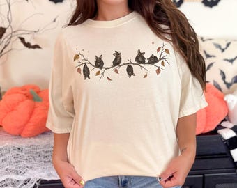 May include: A cream-colored t-shirt featuring a graphic print of five small bats perched on a thin, leafless branch. The illustration includes scattered autumn leaves in shades of orange and brown, along with small star accents above the bats.
