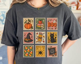 May include: A charcoal gray short-sleeve t-shirt featuring a grid of nine postage stamp-style illustrations. The graphics include a pumpkin, a stack of books with an apple, the text BOO with a ghost, a witch hat, pencils, a black cat, candy corn, a spider with a web, and a chalkboard with a smiley face.