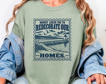 May include: A sage green t-shirt featuring a dark blue graphic design shaped like a postage stamp. The print depicts a lighthouse on a pier with the text "NOBODY ASKED YOU TO REDECORATE OUR HOMES." and the names of the Great Lakes listed below.