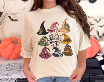 May include: A cream-colored t-shirt featuring a graphic print of eight distinct witch hats in various colors and styles, arranged around the central text "which witch are you?" surrounded by small floral accents and leaves.