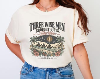 May include: A cream-colored t-shirt featuring a graphic print with the text "THREE WISE MEN BROUGHT GIFTS. Take Notes." and "MATTHEW 2:11". The illustration depicts three figures on camels under a star, framed by palm trees and pink flowers.
