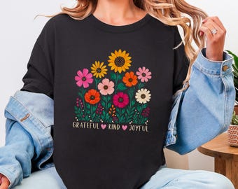 May include: A black t-shirt featuring a colorful graphic of various flowers including sunflowers, daisies, and cosmos in shades of yellow, orange, pink, and red. Below the flowers, the text reads GRATEFUL, KIND, and JOYFUL.