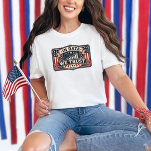 Puede incluir: Camiseta blanca con un gráfico patriótico que dice "IN DATA WE TRUST" en rojo, blanco y azul. La camiseta se combina con vaqueros azules. Una bandera americana se sostiene en la mano izquierda.