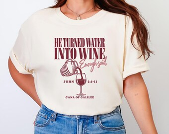 May include: A cream-colored short-sleeve t-shirt featuring dark red text that reads HE TURNED WATER INTO WINE Enough said. JOHN 2:1-11 CANA OF GALILEE. The graphic includes a line drawing of a pitcher pouring liquid into a wine glass.