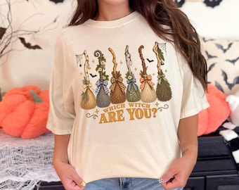May include: A cream-colored short-sleeve t-shirt featuring a graphic of six distinct witch brooms decorated with bows, greenery, and small charms. Below the brooms, the text reads "WHICH WITCH ARE YOU?" in a bold, golden-brown, stylized font.
