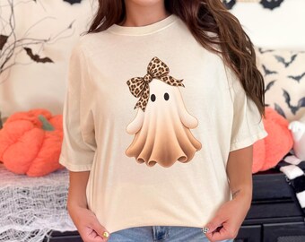May include: A cream-colored short-sleeve t-shirt featuring a central graphic of a ghost with a brown and black leopard print bow. The ghost has two black circular eyes and a light brown gradient body, centered on the front of the garment.