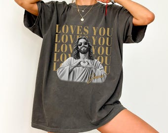 May include: A charcoal gray short-sleeve t-shirt featuring a graphic of a person with long hair and sunglasses making a heart shape with their hands. The text LOVES YOU is repeated in gold behind the figure, with the word Jesus in gold script.