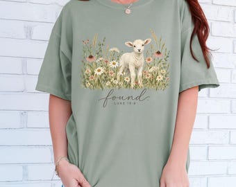 May include: A sage green short-sleeve t-shirt featuring a graphic of a white lamb standing in a field of colorful wildflowers. Below the illustration, the text reads "found." and "LUKE 15:6" in a dark cursive and sans-serif font combination.