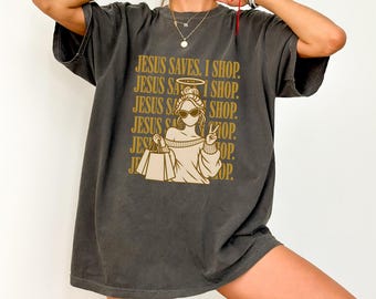 May include: A charcoal gray oversized t-shirt featuring a gold graphic print of a person with a halo, sunglasses, and shopping bags. The text "JESUS SAVES. I SHOP." is repeated in the background behind the central figure in a gold font.