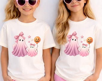 May include: A cream-colored t-shirt featuring a graphic of two pink ghosts holding hands, with the larger ghost wearing a pink bow and the smaller ghost holding an orange jack-o'-lantern balloon, surrounded by small pink stars.