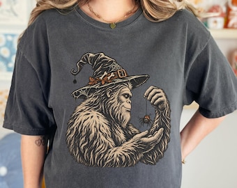 May include: A charcoal gray short-sleeve t-shirt featuring a graphic illustration of a furry creature wearing a pointed witch hat adorned with bones and spiderwebs, holding a small spider dangling from a thread between its fingers.