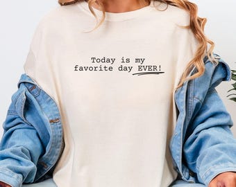 May include: A cream-colored crew neck t-shirt featuring the black printed text Today is my favorite day EVER! with a horizontal line underneath the word EVER. The shirt is worn under a blue denim jacket with long sleeves and visible metal buttons.