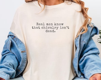 May include: A cream-colored crewneck t-shirt featuring the black printed text "Real men know that chivalry isn't dead." The shirt is worn under a blue denim jacket with long sleeves and visible seams, creating a layered casual appearance.