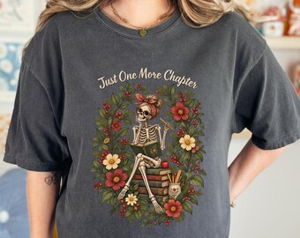 May include: A charcoal gray t-shirt featuring a graphic of a skeleton wearing a red headband, sitting on a stack of books while reading. The design includes the text "Just One More Chapter" surrounded by a wreath of red and white flowers.