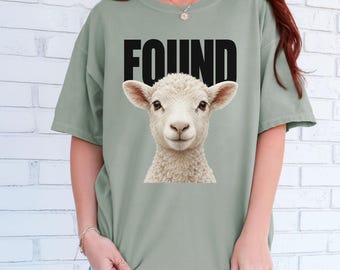 May include: A sage green short-sleeve t-shirt featuring a graphic of a white lamb's head centered on the chest. Above the lamb, the word FOUND is printed in bold, black, capital letters. The shirt has a crew neckline and a relaxed, loose fit.