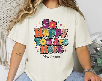 May include: A cream-colored short-sleeve t-shirt featuring a multi-colored graphic print with the text "So Happy You Are Here" and "Mrs. Johnson" below it. The design includes various patterns like polka dots and checkers in orange, blue, and pink.