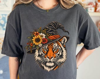 May include: A charcoal gray short-sleeve t-shirt featuring a graphic of a tiger wearing a black witch hat adorned with a cluster of sunflowers, orange blossoms, and green leaves. The tiger has orange, black, and white fur with detailed whiskers.