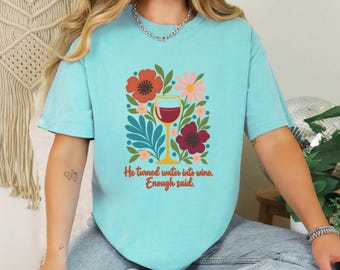 May include: A light blue t-shirt featuring a graphic of a wine glass surrounded by colorful flowers and leaves. The text printed below the illustration reads, "He turned water into wine. Enough said." The shirt has a crew neckline and short sleeves.
