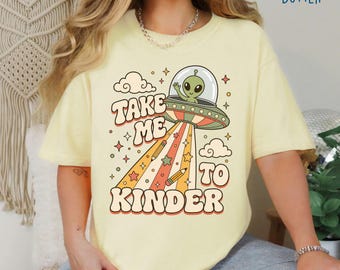 May include: A butter yellow t-shirt featuring a graphic of a green alien in a flying saucer beaming down pencils. The text reads "TAKE ME TO KINDER" in a retro font, surrounded by stars and clouds. The design uses orange, red, and green colors.