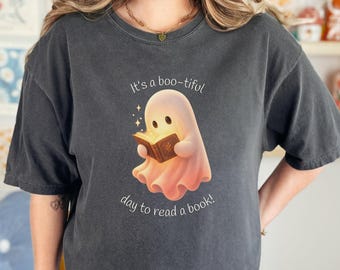 May include: A charcoal gray t-shirt featuring a graphic of a small, light-colored ghost reading an open book that emits a warm glow. The text above the ghost reads It's a boo-tiful and the text below reads day to read a book! The ghost has large eyes.