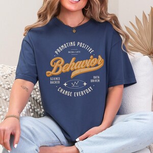 Puede incluir: Una camiseta de manga corta azul marino con el texto "PROMOTING POSITIVE BEHAVIOR" en un diseño circular. El centro muestra "BCBA LIFE" y "Behavior" en una fuente cursiva grande, junto con el texto "SCIENCE BACKED" y "DATA DRIVEN".