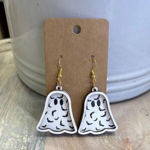 Op de afbeelding: Een paar Halloween-oorbellen. Elke oorbel heeft de vorm van een witte geest met zwarte vleermuis- en oogdetails. De oorbellen hangen aan goudkleurige haken en worden gepresenteerd op een bruine kaart.