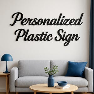 Op de afbeelding: Zwart, cursief geschreven "Personalized Plastic Sign" aan een witte muur. Daaronder een grijze bank met een blauw kussen, een houten bijzettafel met een blauwe lamp en een ronde salontafel met een blauwe vaas en kom.