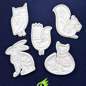 May include: A set of five white plastic cookie cutters featuring embossed designs of an owl, a bird, a squirrel, a rabbit, and a fox. Each cutter displays intricate floral and geometric patterns on the surface, arranged on a dark blue background.