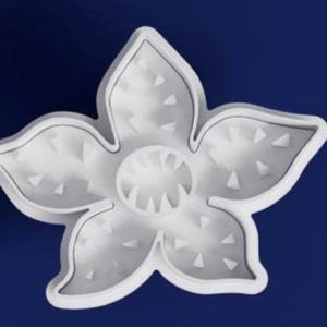 Pode incluir: Um cortador de biscoitos branco em forma de flor de cinco pétalas com uma superfície texturizada. O cortador tem um design circular central e está sobre um fundo azul escuro. As pétalas têm reentrâncias triangulares.
