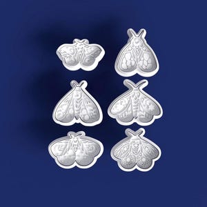 Pode incluir: Seis cortadores de biscoitos brancos em forma de mariposa com desenhos intrincados, dispostos sobre um fundo azul escuro. Os cortadores apresentam padrões detalhados de asas e corpos, perfeitos para projetos de panificação ou artesanato. Os desenhos incluem luas crescentes e padrões florais.