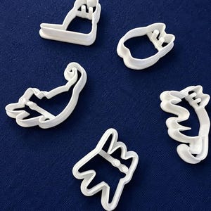 May include: A set of five white plastic cookie cutters, each shaped like a cat in various poses, including a sitting cat, a curled cat, a stretching cat, a cat with a raised tail, and a cat head, arranged on a dark blue textured surface.