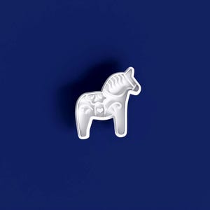 May include: A white Dala horse figurine with detailed floral and heart patterns. The horse is set against a deep blue backdrop, highlighting its intricate design. This decorative item is a classic Scandinavian folk art symbol.