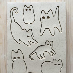May include: A rectangular, off-white ceramic tile featuring seven cat-shaped cutouts. The cats are in various poses, including standing, sitting, and stretching. The tile has a speckled texture and is likely used for crafting or decoration.