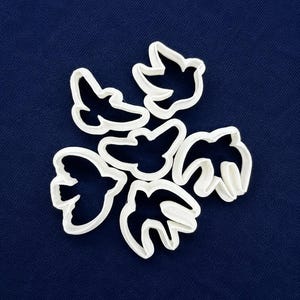 Puede incluir: Un conjunto de cinco cortadores de galletas de plástico blanco con forma de pájaro dispuestos en un grupo. Cada cortador presenta el contorno distintivo de un pájaro en vuelo con las alas extendidas, diseñado para cortar masa en formas.