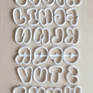 Puede incluir: Cortadores de galletas de plástico blanco con forma de alfabeto en estilo burbuja. Las letras están dispuestas sobre una superficie de madera clara. Un gráfico verde y amarillo se encuentra en la esquina inferior derecha.