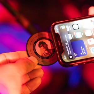 May include: A bronze-coloured NFC tag with a portrait is held near a smartphone. The phone displays a website notification. The tag has the text "ZATO-2005-M" and other text around the edge. The phone shows the time as 7:42.