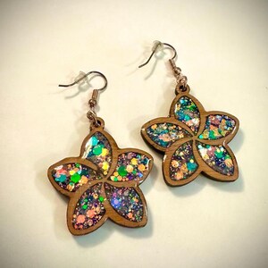 May include: A pair of wooden flower-shaped earrings with a glitter-filled center. The earrings have a light brown wooden frame and are filled with colorful glitter in shades of blue, green, pink, and purple. The earrings have copper-colored ear wires.