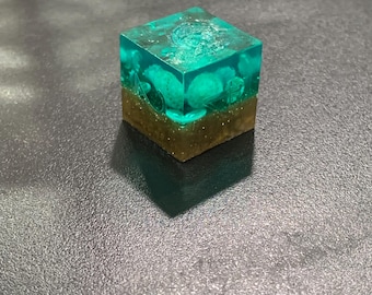 May include: A clear resin cube with a teal interior, featuring small, light-colored stones and a sandy-brown base. The cube is sitting on a dark surface, casting a shadow. The overall effect is a miniature, encapsulated landscape.