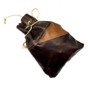 May include: A handcrafted leather pouch in shades of brown. The bag features a drawstring closure with a natural twine cord. The top portion is a darker, textured leather, with a lighter brown patch sewn on the side. The bottom is a smooth, deep brown.